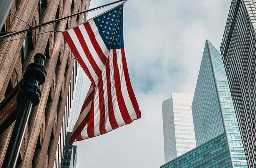 The USA or united states of america flag on a flagpole near skyscrapers under a cloudy sky